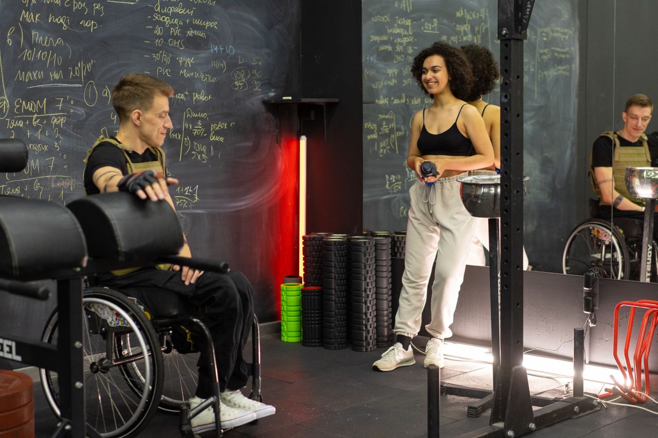 Home Two people in a gym, one in a wheelchair and one standing, preparing for a workout.